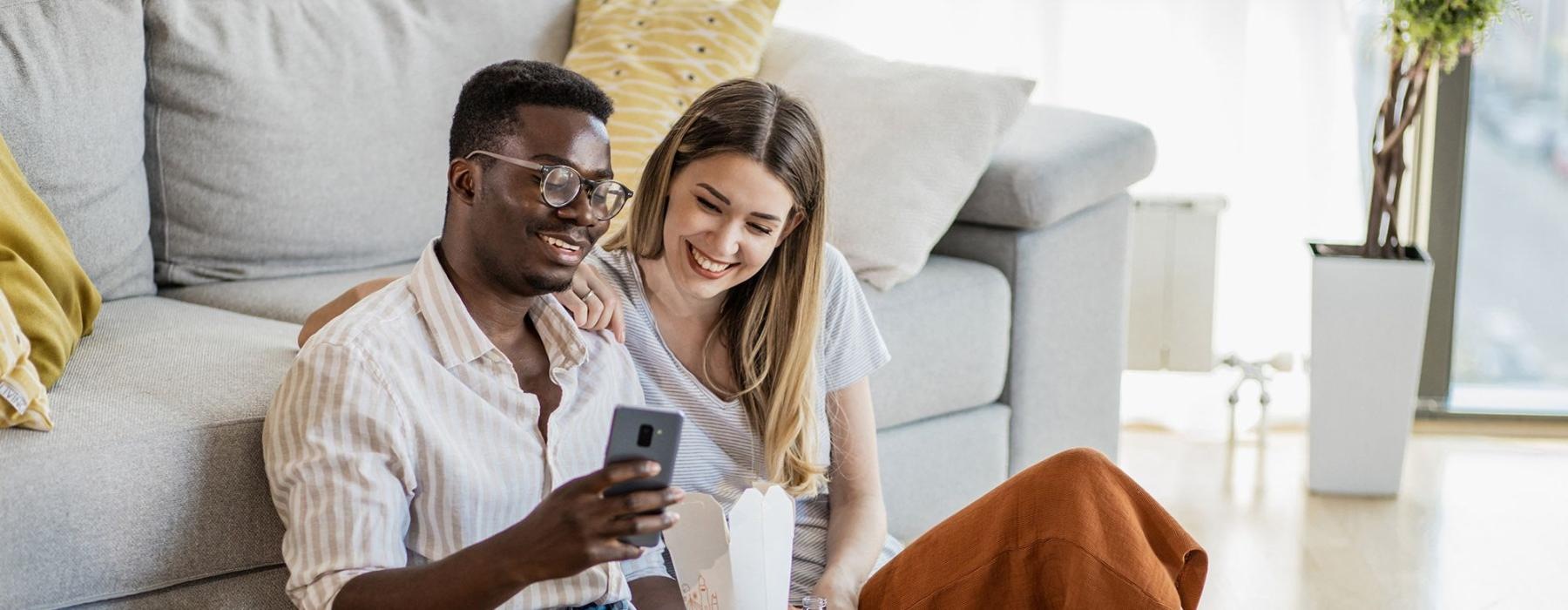 a man and woman with take out, sit against a couch on their living room floor and watch at their cell phone
